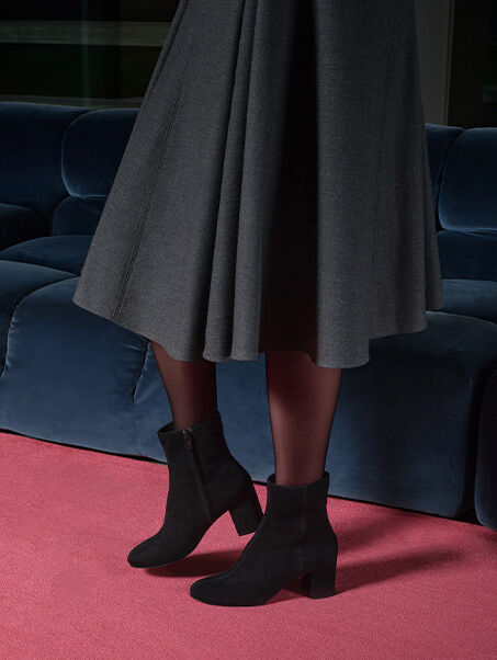 Woman in front of a blue sofa wearing a pleated skirt, tights and ankle boots, while low-heeled pumps rest on pink carpet | GEOX | GEOX